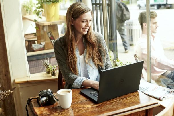 Young woman sitting at a table in a cafe with a cup of coffee and a digital camera, looking out the window, using a Dell Inspiron 14 3000 Series (Model 3451 Iris) notebook computer.