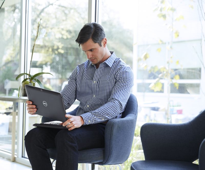 Man in professional clothing seated on the ground floor of an office with large, open windows and a Dell Latitude 13 7000 Series (Model 7350), 13 inch 2-in-1 notebook computer in his lap. He is holding the tablet screen slightly above the keyboard.